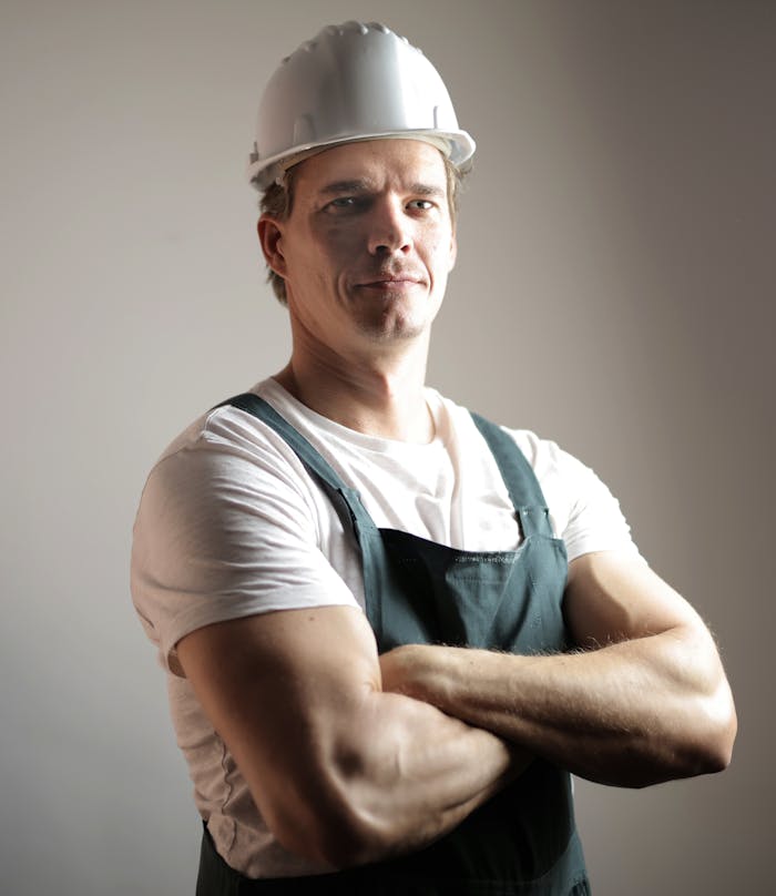 Positive confident constructor in uniform and white hardhat standing with arms crossed and looking at camera against gray background